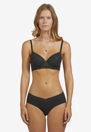 Frau steht frontal und trägt einen schwarzen Spitzen-BH und schwarze Bikini-Unterwäsche vor einem schlichten weißen Hintergrund.