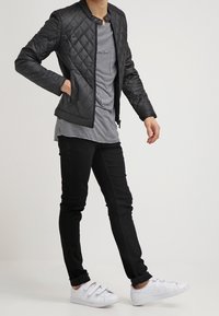 Person som bär en svart vadderad jacka, randig skjorta, svarta byxor och vita sneakers med kardborre som står mot en enfärgad bakgrund.