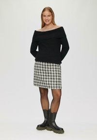 Schwarzer, schulterfreier Strickpullover, Rock im Hahnentrittmuster in Schwarz und Weiß, und schwarze Stiefeletten mit dicken grünen Sohlen.