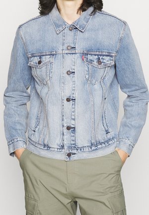 Personne portant une veste en jean bleu clair boutonnée, les mains dans les poches d’un pantalon cargo vert olive, montrée du cou aux hanches.