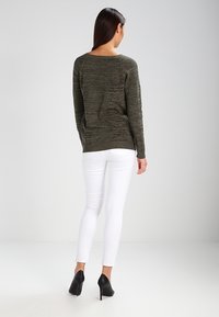 Olivgrön stickad tröja med avslappnad passform, ribbat halslinne och långa ärmar, kombinerad med vita skinny jeans och svarta högklackade skor.