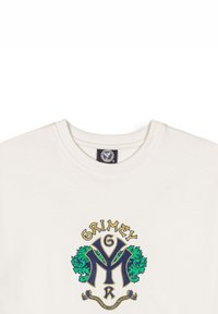 Camiseta de algodón color beige claro con un gráfico bordado colorido que presenta motivos de león verde, texto amarillo y azul, y cuello redondo.
