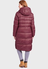 Bordeauxfarbene Steppjacke mit Kapuze, gestepptem Design und langer Länge. Verfügt über seitliche Schlitze am Saum und wird über einer hellen Jeans und Stiefeln getragen.