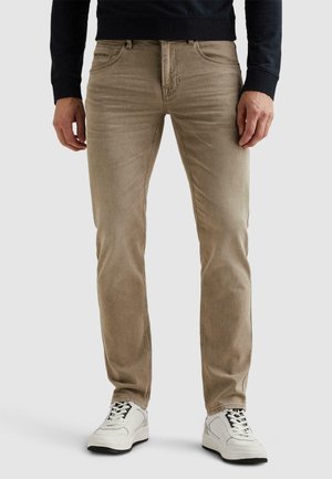 Hombre con jeans ajustados beige y zapatillas blancas, de pie frente a un fondo liso de color gris claro.