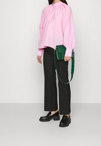 Blusa rosa a collo alto con maniche voluminose, abbinata a pantaloni neri a gamba larga e mocassini neri di grandi dimensioni. Borsa verde con motivo coccodrillo.