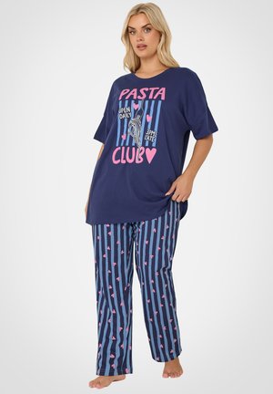 Lyshåret kvinde iført navyblå oversized T-shirt med teksten "Pasta Club" og stribede pyjamasbukser med lyserøde hjerter, stående barfodet.