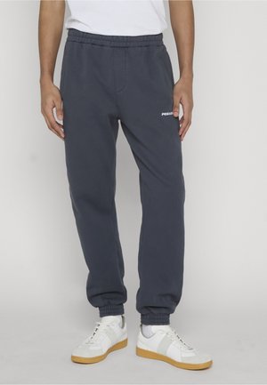 Osoba nosząca ciemnoszare spodnie dresowe z elastycznymi ściągaczami i białe buty sportowe z gumowymi podeszwami, stojąca na jednolitym tle.