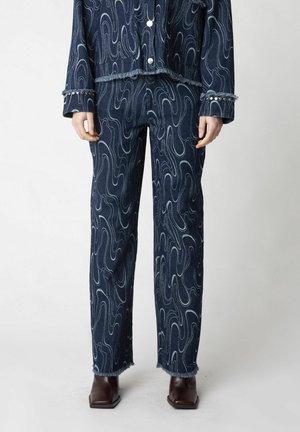 Person iført mørkeblå denimbukser og matchende jakke med lyse swirl mønstre, frynser på kanterne og brune støvler med firkantet tå.