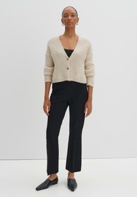 Cardigan beige côtelé avec des manches trois-quarts, un décolleté en V et une fermeture à bouton unique. Associé à un pantalon noir ajusté et des ballerines noires pointues.