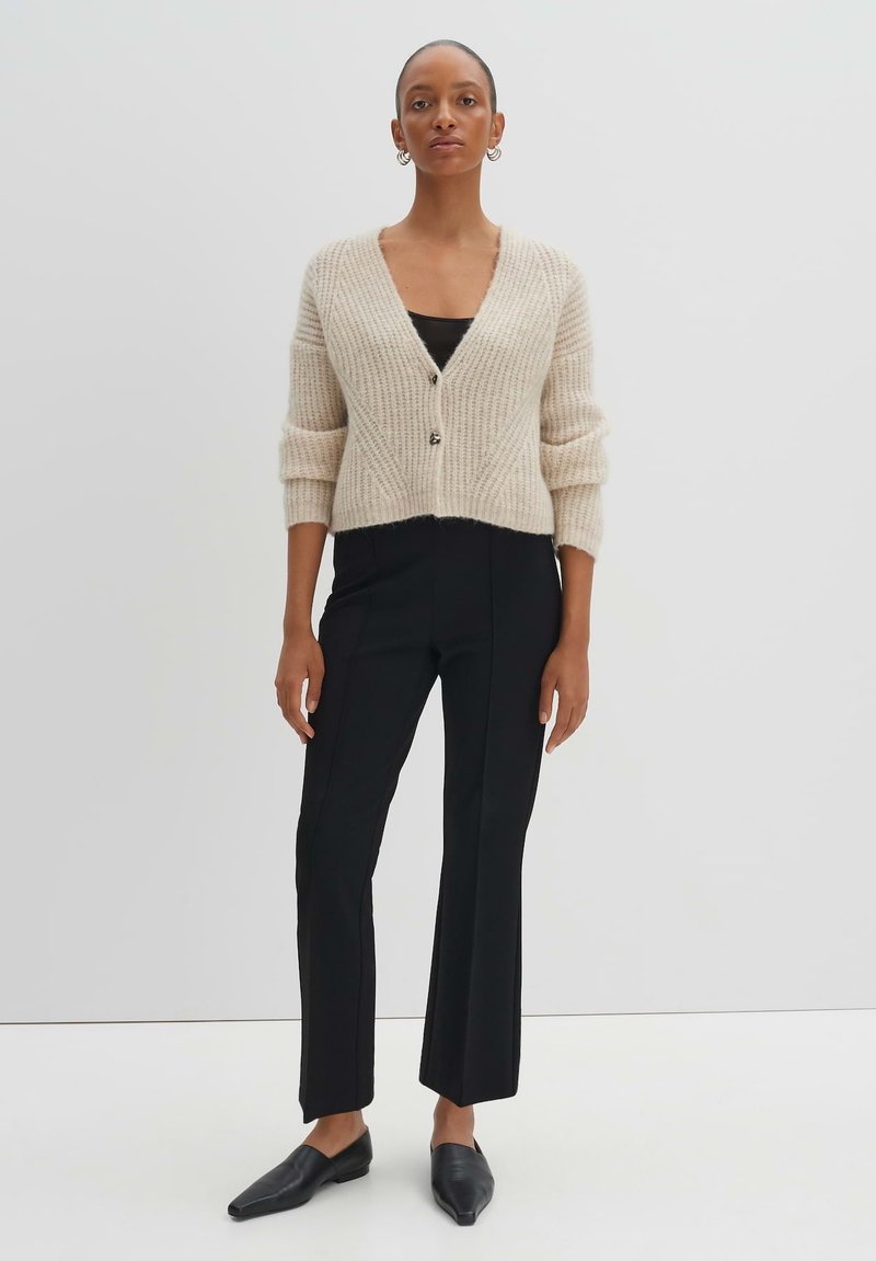 Beige Rippstrick-Cardigan mit Dreiviertelärmeln, V-Ausschnitt und einer einzelnen Knopfleiste. Getragen mit schwarzen, figurbetonten Hosen und spitzen schwarzen Ballerinas.