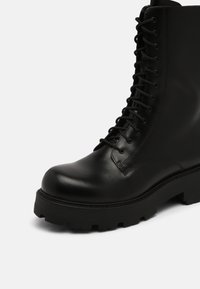 Bottine en cuir noir avec bout rond, dotée d'un laçage et d'une semelle épaisse texturée avec des motifs de crampons profonds.