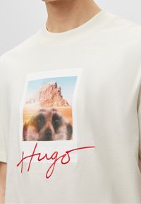 Camiseta de algodón color crema con un estampado gráfico de un paisaje desértico y un primer plano de la cara de un animal; texto bordado en rojo "Hugo".