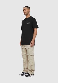 Sort kortærmet t-shirt med et logo, parret med beige cargo bukser med flere lommer og en løs pasform, suppleret med sort-hvide sneakers.