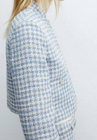 Donna con indosso un giubbotto in tweed a quadri blu e bianchi e una gonna coordinata, in piedi contro uno sfondo chiaro e uniforme.