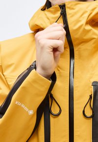 Χέρι που τραβάει το μαύρο φερμουάρ σε κίτρινο μπουφάν Gore-Tex με ρυθμιζόμενο κουκούλα και μαύρες λεπτομέρειες.