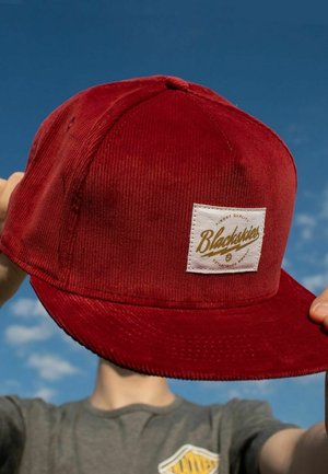 Red corduroy baseball cap with a white patch displaying "Blackskies" in gold script. Features a curved visor and textured fabric.