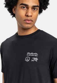 Černé tričko s krátkým rukávem vyrobené z texturované látky. Obsahuje bílé logo Quiksilver a grafické návrhy: znak míru, rybu a stylizovanou vlnu.