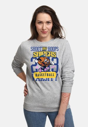 Kobieta z długimi brązowymi włosami, ubrana w szarą bluzę z postacią z kreskówki koszykarza oraz tekstem o koszykówce NY hoops varsity.