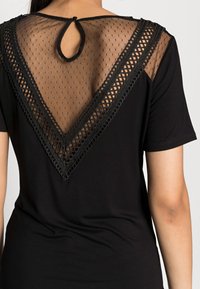 Kobieta nosząca czarny top z krótkim rękawem z przezroczystej tkaniny w kropki oraz dekoracyjną koronk�ą tworzącą kształt litery V na górnej części pleców, z zapięciem na guziczek w kształcie otworu.
