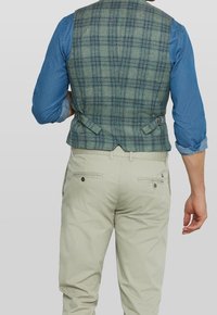 Grön rutig väst med en figursydd form, med detaljer i ryggen som har dubbelspänne, bärs över en blå långärmad skjorta och ljusa byxor.