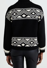 Svart stickad tröja med ett geometriskt vitt mönster, polokrage och långa ärmar med randiga detaljer vid muddar och nederkant.