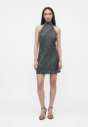 Donna che indossa un vestito corto senza maniche, con collo alto e righe diagonali verdi, blu e nere, abbinato a sandali neri con tacco alto.