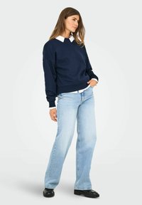 Felpa blu navy con scollo ampio, abbinata a una camicia bianca con colletto sottostante. Un paio di jeans azzurri e scarpe nere completano l'outfit.