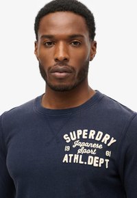 Navy blue sweatshirt made of cotton with a crew neckline, featuring an embroidered logo in cream color on the front and ribbed cuffs.