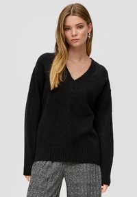 Schwarzer V-Ausschnitt Pullover mit weiten Ärmeln und geripptem Saum. Weiche Textur, minimalistisches Design, kombiniert mit gemusterten grauen Hosen.