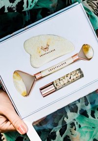 Set di skincare in cristallo con un rullo di giada, un attrezzo gua sha e un vial di cristalli. Gli strumenti presentano finiture in oro rosa e superfici lisce di colore chiaro.
