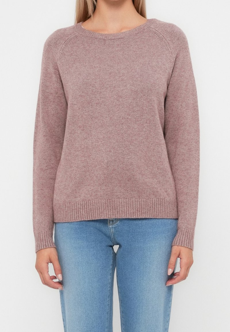 Femme portant un pull tricoté mauve à col rond et un jean bleu clair, debout, les bras détendus de chaque côté, sur un fond uni.