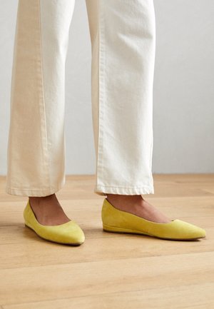 Personne portant un pantalon ample écru et des chaussures plates en suède jaune, debout sur un sol en bois clair.