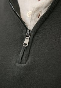 Mörkgrå tröja med hög krage, utrustad med en silverdragkedja och strukturerat stickat mönster. Vit skjortkrage som syns under.