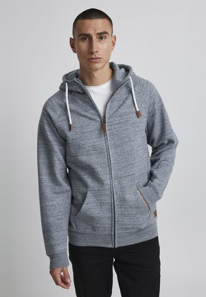 Haut à zip gris avec une finition texturée, comportant une capuche à cordon, deux poches avant et des accents en cuir marron.