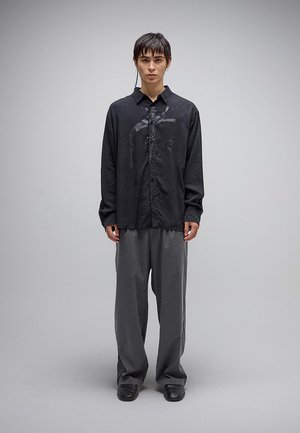 Νεαρός άνδρας που φοράει άνετο μαύρο πουκάμισο με κουμπιά και διακριτικό σχέδιο, γκρι παντελόνι φαρδιάς γραμμής και μαύρα παπούτσια, στέκεται μπροστά σε απλό φόντο.