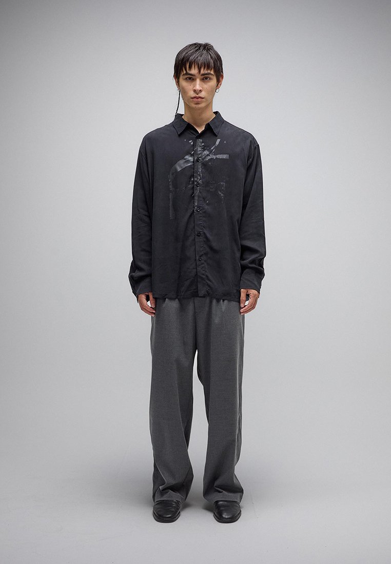 Νεαρός άνδρας που φοράει άνετο μαύρο πουκάμισο με κουμπιά και διακριτικό σχέδιο, γκρι παντελόνι φαρδιάς γραμμής και μαύρα παπούτσια, στέκεται μπροστά σε απλό φόντο.