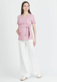 T-shirt de maternité en tissu rose doux, avec un détail de nouage sur le côté, un décolleté rond et des manches courtes. Associé à un pantalon large blanc.