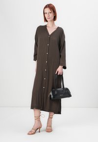 Femme portant une robe longue marron à boutons, tenant un sac à main noir, se tenant sur un fond blanc avec des sandales à talons à lanières beiges.