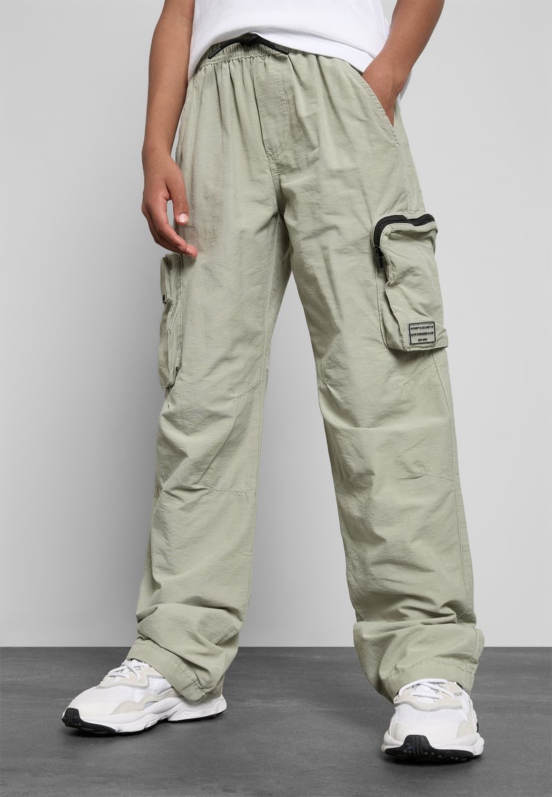 Personne portant un pantalon cargo vert clair avec des poches latérales et des baskets blanches, debout contre un mur et un sol gris.