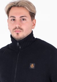 Felpa nera con zip, dotata di un collo alto a coste, caratterizzata da un piccolo emblema arancione del marchio sul lato sinistro del petto. Texture a maglia liscia.