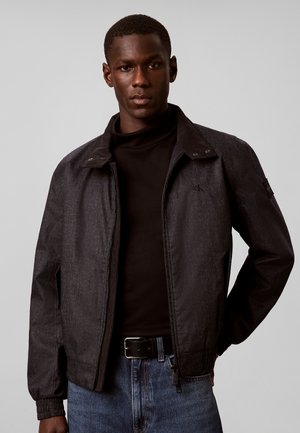 Mann trägt dunkelgraue Zip-Up-Jacke, schwarzen Rollkragenpullover und blaue Jeans und steht vor hellgrauem Hintergrund.