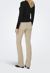 Femme debout de dos, portant un pantalon beige en velours côtelé, un haut noir à manches longues et des chaussures noires devant un fond clair uni.