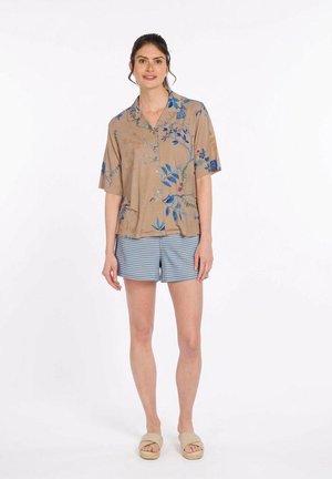 Woman wearing a tan short-sleeve shirt with blue floral print, blue striped shorts, and beige slippers, standing against a white background.