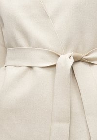 Μπεζ παλτό τύπου wrap από απαλό ύφασμα, με ζώνη δεμένη στη μέση και λεία υφή, με επίπεδες ραφές και χωρίς ορατά κλείστρα.