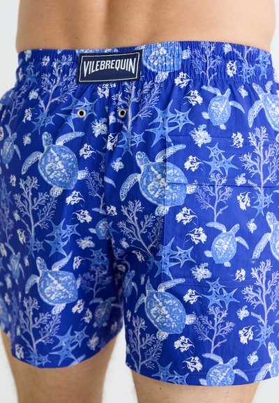 Costum de baie bleu cu un model de țestoasă și corali. Dispune de un brâu elastic cu un eticheta de marcă și două orificii pentru ventilație.