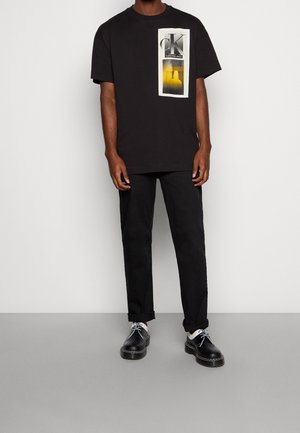 Musta puuvillapaita, jossa on graafinen printti "CK"-logolla. Yhdistetty mustiin housuihin ja mustiin kenkiin, joissa on valkoisia yksityiskohtia ja nauhoja.