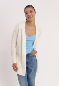 White ribbed cardigan with open front, two pockets; layered over a light blue tank top. Jeans feature a classic cut with slight fading.