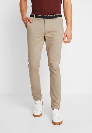 Mężczyzna w beżowych obcisłych spodniach, czarnym pasku, białych trampkach i ciemnoczerwonej koszuli, stojący z jedną ręką w kieszeni.