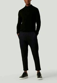 Schwarzer Rollkragenpulli, kombiniert mit dunklen Hosen, die einen schmalen Schnitt und Taschen aufweisen, ergänzt durch schwarze Sneaker mit weißen Sohlen.