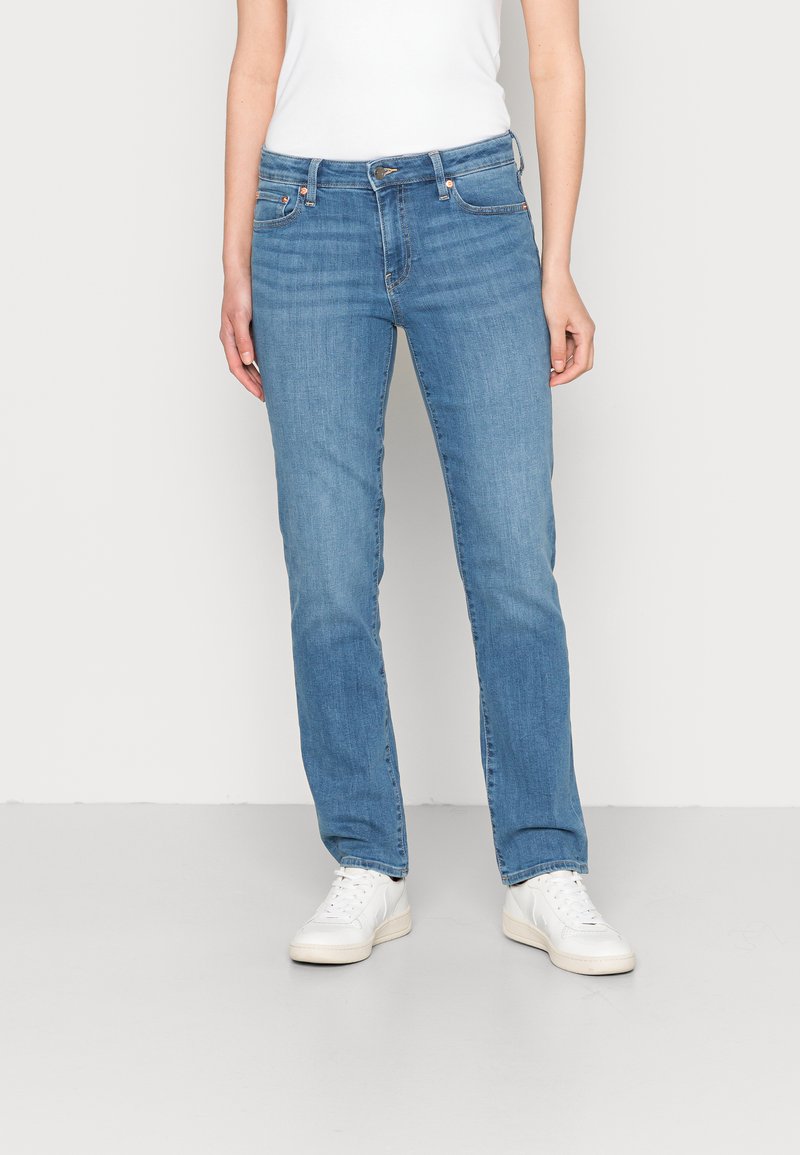 Jeans de ganga azul claro com um design de perna reta, cinco bolsos e acabamentos em metal. Usados com uma t-shirt branca e ténis brancos.